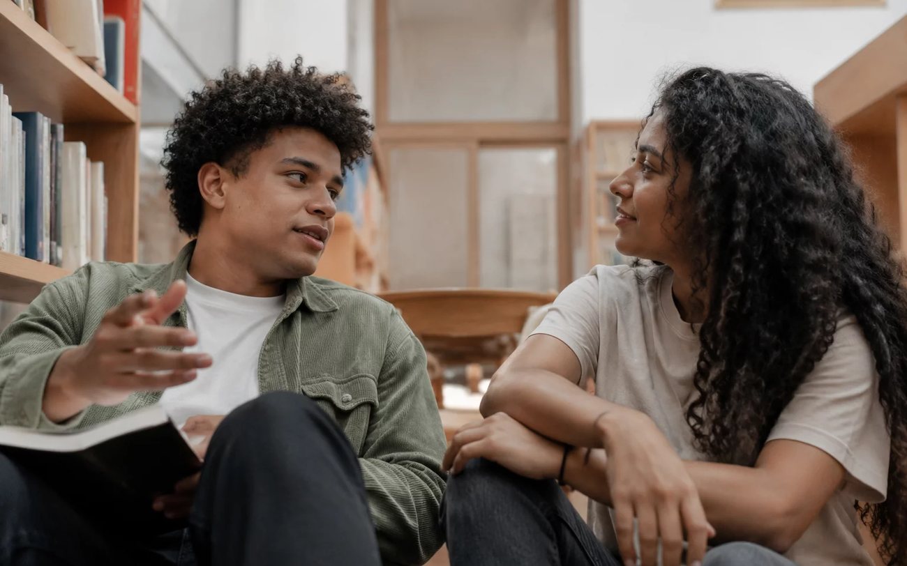 Três pessoas sentadas a uma mesa comprida, no meio de uma conversa numa sala de janelas altas