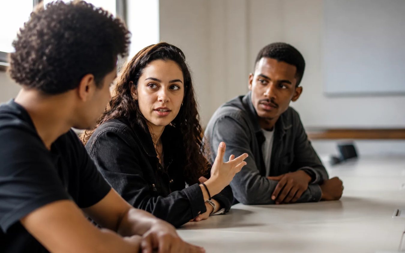 Três participantes conversam ao redor de uma mesa comprida numa sala clara de janelas altas